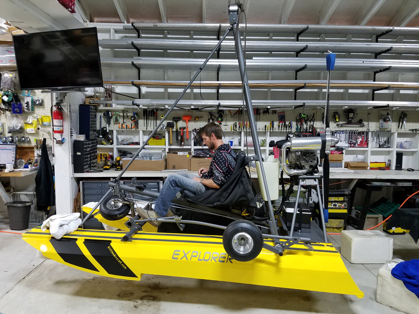 Yellow ultralight amphibious aircraft boat labeled 'Explorer' in a workshop setting with tools and equipment.